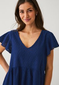Navyblaues V-Ausschnittkleid mit kurzen, gerüschten Ärmeln, das über eine florale Spitzenstruktur und einen ausladenden Saum verfügt, was eine entspannte Silhouette schafft.
