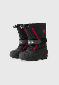 Botas de nieve negras con parte superior de nylon acolchada, acentos rojos, una correa gris con el texto "SOREL" y una suela de goma texturizada para mayor agarre.