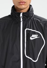 Svart Nike vindjacka med hög krage, vit dragkedja och diagonalt logotypakcent. Funktioner en ficka på bröstet med dragkedja. Slät textur.