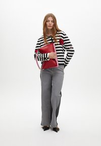 Czerwono-różowa kopertówka o teksturowanym wykończeniu, trzymana przez modelkę w czarno-białym swetrze w paski z haftowanymi detalami i szarych dżinsach.