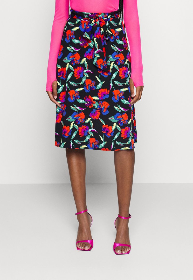 Jupe noire longueur genou à imprimé floral rouge et violet, taille nouée, associée à un haut rose à manches longues et des talons ouverts roses.