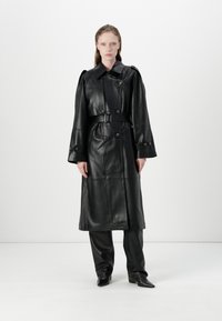 Black leather trench coat with a belted waist, large buttons, and wide sleeves. Worn over matching black pants and pointed boots.