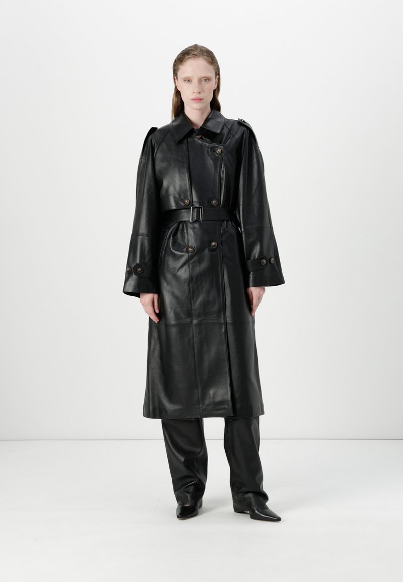 Black leather trench coat with a belted waist, large buttons, and wide sleeves. Worn over matching black pants and pointed boots.