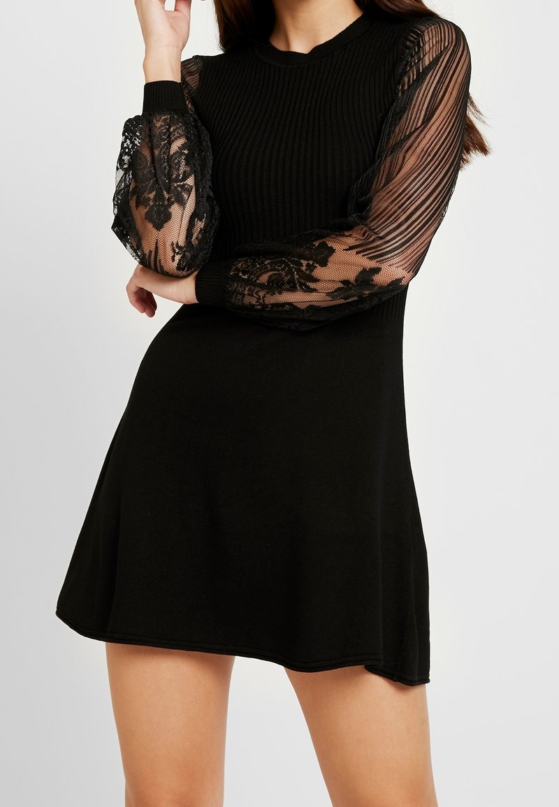 Femme portant une robe noire avec un corsage côtelé et des manches longues en dentelle transparente, debout les bras croisés à la taille.