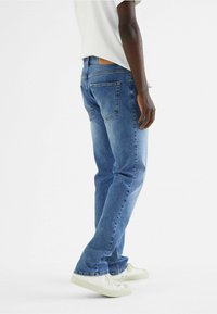 Blauwe spijkerbroeken met een relaxed fit, vijf zakken, standaard stiksel en een tan leren patch op de achterkant van de tailleband. Gecombineerd met witte schoenen.