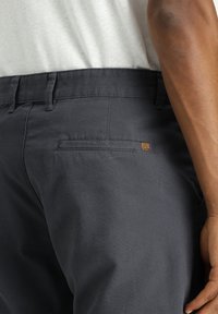 Pantalones gris oscuro de tela suave, con un bolsillo trasero que presenta un pequeño acento de cuero marrón y una cinturilla estándar.