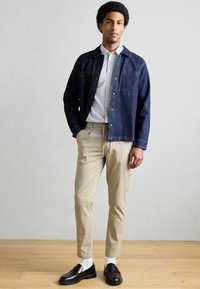 Σκούρο τζιν πουκάμισο, πουκάμισο με ρίγες από κάτω, μπεζ παντελόνι, μαύρα μοκασίνια και λευκές κάλτσες· ορατά κουμπιά και λεπτομέρεια τσέπης.