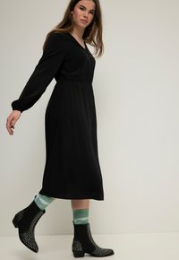 Vestido negro de manga larga con cintura ajustada y falda fluida. Llevado con botas de tobillo negras con tachuelas y calcetines a rayas verdes.