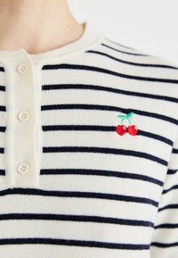 Stripet bomull henley med kremfarget bakgrunn og marineblå striper, med tre knapper og en liten rød kirsebærbroderi detalj.