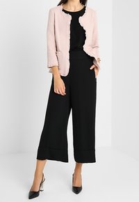 Chaqueta texturizada de color rosa claro con bordes con volantes, combinada con una blusa negra y pantalones anchos negros, finalizada con tacones altos negros.