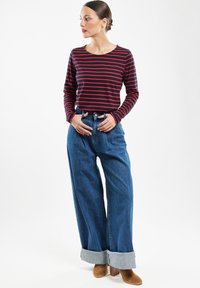 Røde og marineblå stribede langærmede top sammen med højwaist, brede ben blå jeans med opsmøg og brune hæle støvler. Neutral baggrund.