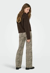 Frau mit langen Haaren, die einen dunkelbraunen Pullover, weit geschnittene Hose mit Leopardenmuster und schwarze Schuhe trägt, steht vor einem schlichten hellen Hintergrund.