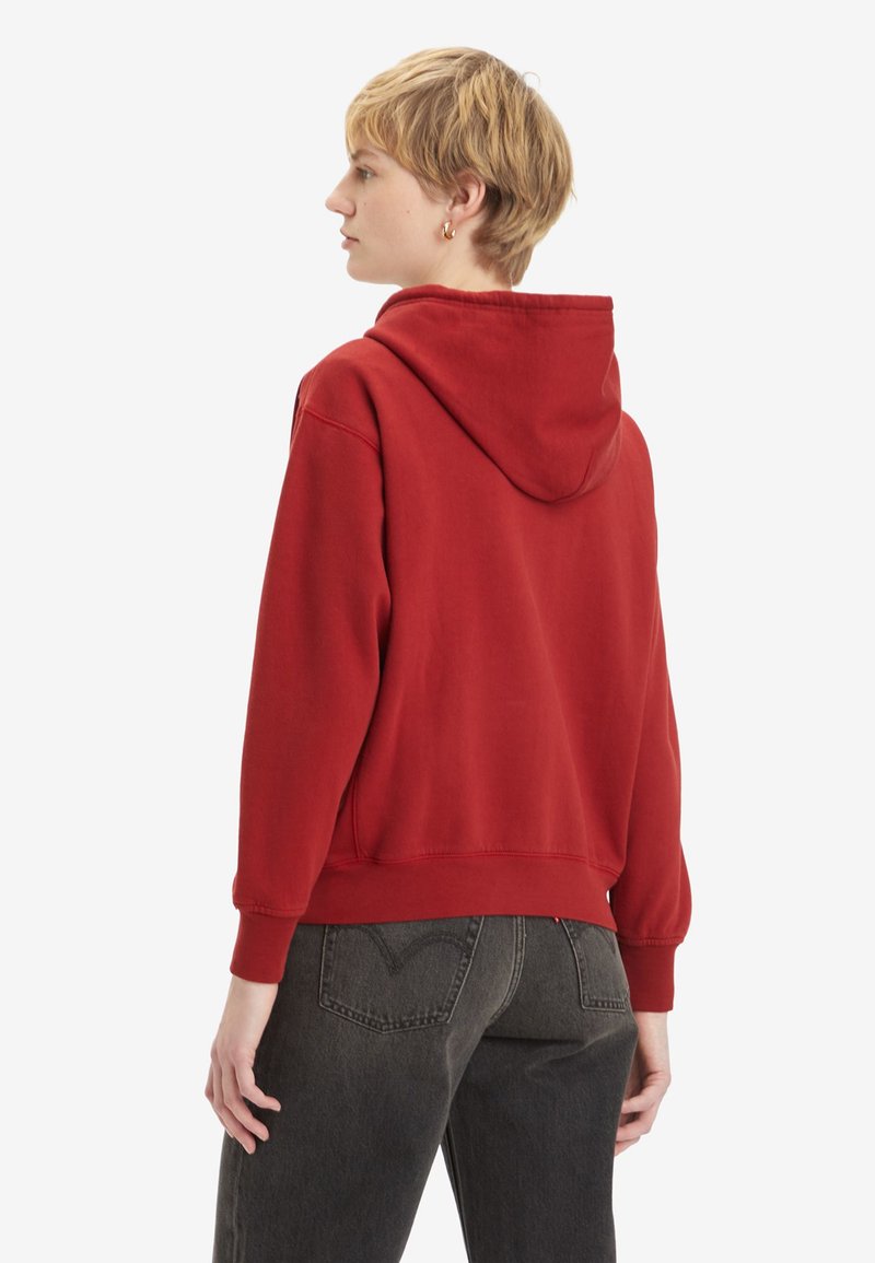 Sweatshirt rouge à capuche en tissu texturé, avec une coupe décontractée et des poignets côtelés. Associé à un jean noir taille haute, affichant un style décontracté.