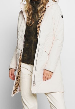 Femme portant un manteau d'hiver rose clair avec une doublure imprimé léopard, un pull à fermeture éclair vert foncé et un pantalon clair.