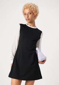 Abito nero con un corpo testurizzato e maniche a pois bianchi; include una borsa lavanda con un design triangolare e manico annodato.