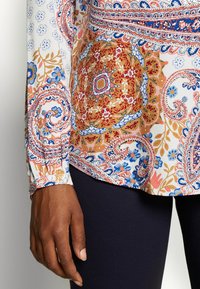 Blouse à manches longues en tissu léger, présentant un motif cachemire coloré en rouges, bleus et ors, avec un détail de poignets froncés.