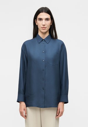 Kobieta z długimi ciemnymi włosami, ubrana w zapinaną na guziki granatową koszulę z długim rękawem i beżowe spodnie, stojąca na tle jednolitego jasnego tła.