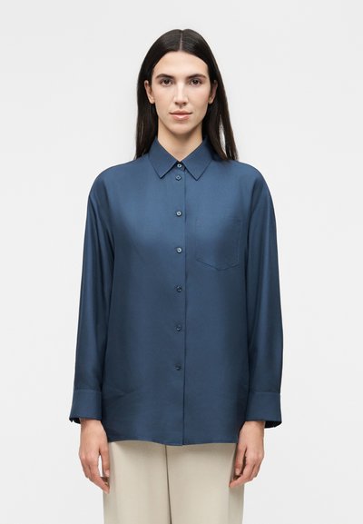 Femme aux longs cheveux foncés portant une chemise bleu marine boutonnée à manches longues et un pantalon beige, debout devant un fond clair uni.
