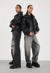 To personer står rygg mot rygg iført svarte skinnjakker, løse falmede jeans og svarte kraftige sko mot en ensfarget bakgrunn.
