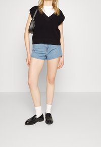 Top negro sin mangas de punto con escote en V, combinado con shorts de mezclilla azul claro. La modelo lleva calcetines blancos y mocasines negros.