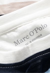 Étiquette en tissu blanche avec un texte gris indiquant "Marc O'Polo EST. À STOCKHOLM." Bords coupés visibles, avec des tissus bleu marine et beige en dessous.
