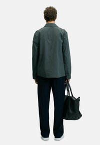 Donkergrijze lichtgewicht jas met lange mouwen, gecombineerd met marineblauwe broek en witte sneakers. Het model houdt een zwarte leren tas vast.