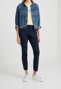 Jeansjacka i medelblått med en klassisk passform, bärs över en gul t-shirt med en blå logga, i kombination med marinblå slim-fit byxor och vita sneakers.