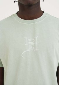 Camiseta de algodón verde claro con cuello redondo y logo impreso en blanco que presenta letras estilizadas en la parte delantera. Textura suave y lisa.