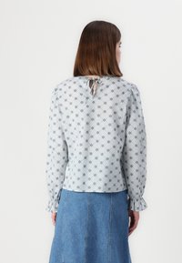 Blusa azul clara com um padrão floral, mangas longas, punhos franzidos e costas recortadas com um fecho de laço; combinada com uma saia de jeans.