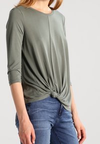 Grünes Shirt mit langen Ärmeln und geknotetem Vorderdesign, aus weichem Stoff, kombiniert mit blauen Jeans.