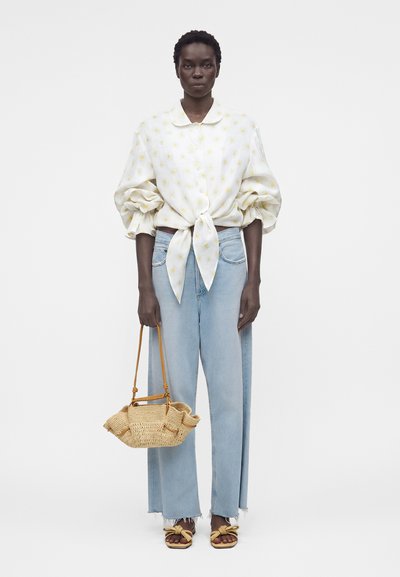 Γυναίκα που φοράει μια λευκή μπλούζα με φουσκωτά μανίκια και κίτρινα πουά, ανοιχτόχρωμα μπλε τζιν, δεμένη ψάθινη τσάντα και κίτρινα σανδάλια με κόμπο.
