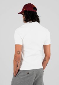 Mężczyzna z kręconymi włosami, noszący bordową czapkę, białą koszulę z krótkim rękawem i szare spodnie, z tatuażami na obu ramionach, stojący tyłem.