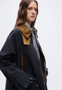 Γυναίκα που φοράει μπλε σκούρο σακάκι με καφέ γιακά και διακοσμήσεις από βελούδο πάνω από πουκάμισο με ασπρόμαυρες ρίγες και κουμπιά.