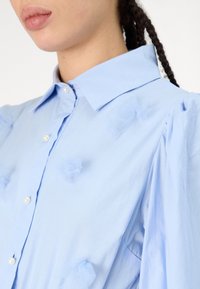 Ανοιχτό μπλε πουκάμισο με κοφτό γιακά, μαργαριταρένια κουμπιά και διακοσμήσεις από υφασμάτινα λουλούδια με υφή τούλι. Έχει φουσκωτά μανίκια και λείο ύφασμα.