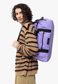 Zaino viola con un design rettangolare, caratterizzato da spallacci neri e una tasca con zip. Indossato da un modello in un maglione a righe.