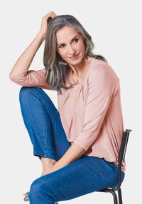 Helles pinkes Oberteil mit Rundhalsausschnitt und Dreiviertelärmeln, kombiniert mit blauen Jeans. Sitzt auf einem schwarzen Stuhl und zeigt eine entspannte Haltung.