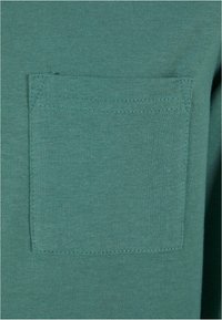 Grünes Baumwoll-T-Shirt mit Rundhalsausschnitt und einer Brusttasche. Der Stoff ist glatt und gleichmäßig in der Textur und weist eine ordentliche Nahtführung an den Kanten auf.