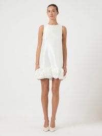 Γυναίκα ντυμένη με ένα αμάνικο λευκό σατέν μίνι φόρεμα με μεγάλες ροζέτες από ύφασμα κατά μήκος του τελειώματος, συνδυασμένο με λευκά μυτερά ψηλοτάκουνα.