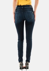 Jean skinny en denim foncé, avec une taille haute, poches arrière avec des rabats pointus, texture lisse et détails de couture subtils.