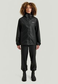 Musta vedenpitävä takki, jossa on korkea kaulus, joustavat hihansuut ja vetoketjusuljin, yhdistettynä mustiin housuihin ja nilkkureihin.