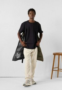 Jonge man in een zwart T-shirt, crème joggingsbroek, beige sneakers, met een donkere pufferjack in zijn hand, staand naast een houten kruk tegen een witte muur.