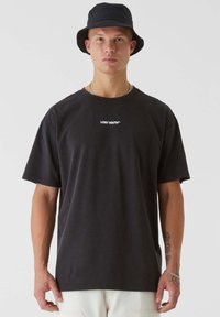 T-shirt en coton noir avec col rond et manches courtes, présentant le logo "LOST YOUTH™" en blanc sur la poitrine. Le modèle porte un chapeau seau assorti.