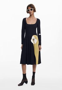 Musta, pitkähihaisen mekon pyöreällä pääntiellä, A-linjaisella mallilla, keltaisella abstraktilla kuviolla sivulla ja teksturoidulla kankaalla.