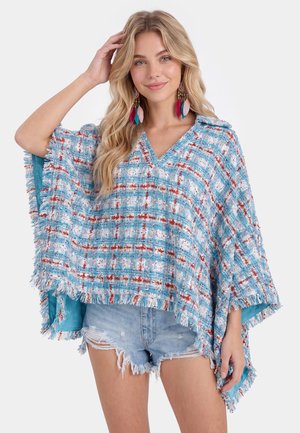 Donna bionda che indossa grandi orecchini colorati di piume, un poncho blu con motivi e frange, e shorts in denim usurati, sorridente con la mano sulla testa.