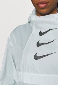 Chaqueta verde menta ligera y translúcida con cuello alto, que presenta tres logotipos de swoosh negros de Nike dispuestos en diagonal en el frente.