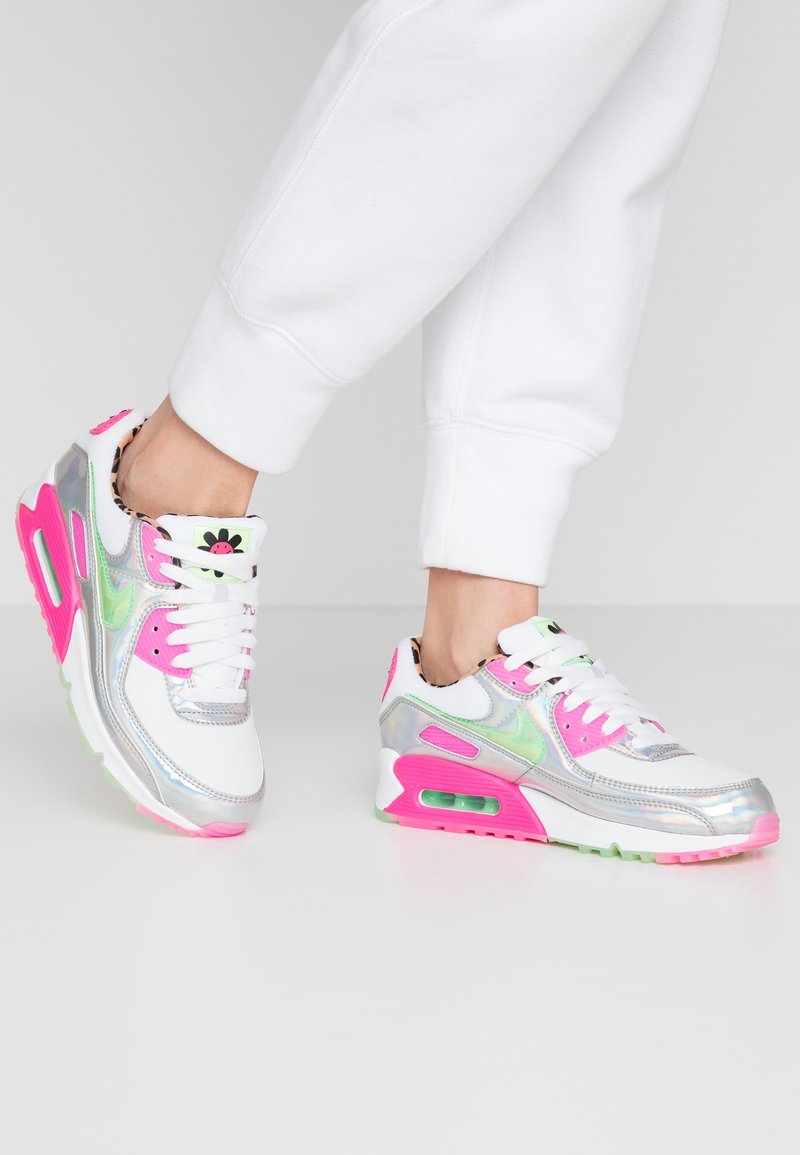 Bílé tenisky s iridescentními akcenty, růžovými detaily a zelenými podrážkami. Obsahují šněrování se viditelným vzduchovým polstrováním.