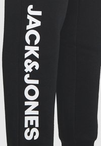 Schwarze Jogginghosen mit einer weichen Textur zeigen großen weißen Text "JACK & JONES" entlang der Seite. Einfaches Design, enganliegende Beinöffnungen.