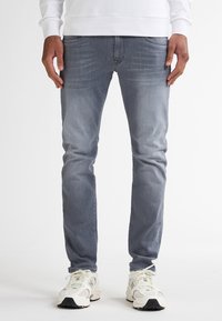 Jeans din denim gri cu o croială slim, având o decolorare subtilă, design clasic cu cinci buzunare, asortați cu adidași albi. 