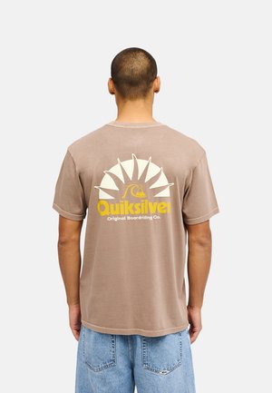 Mężczyzna ubrany w brązowy t-shirt Quiksilver z żółtym logo słońca i fali oraz jasnoniebieskie denimowe szorty, widziany od tyłu.