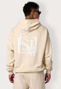 Haut à capuche couleur crème avec un grand logo géométrique blanc au dos. Fabriqué en matériau doux, il présente une coupe décontractée et une capuche avec cordon de serrage.